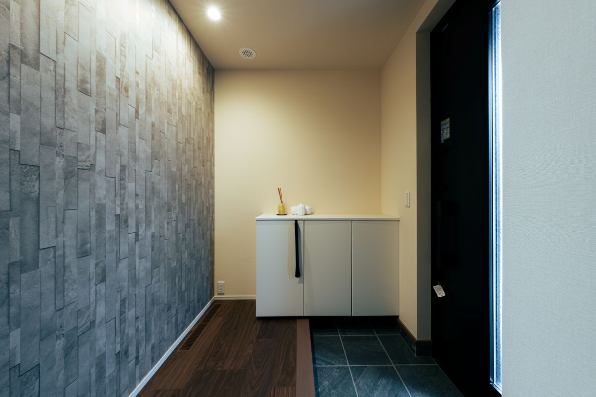 モダンな玄関ホールの内装。壁一面にグレーの石目調アクセントクロスが貼られ、白いシューズボックスが置かれている。