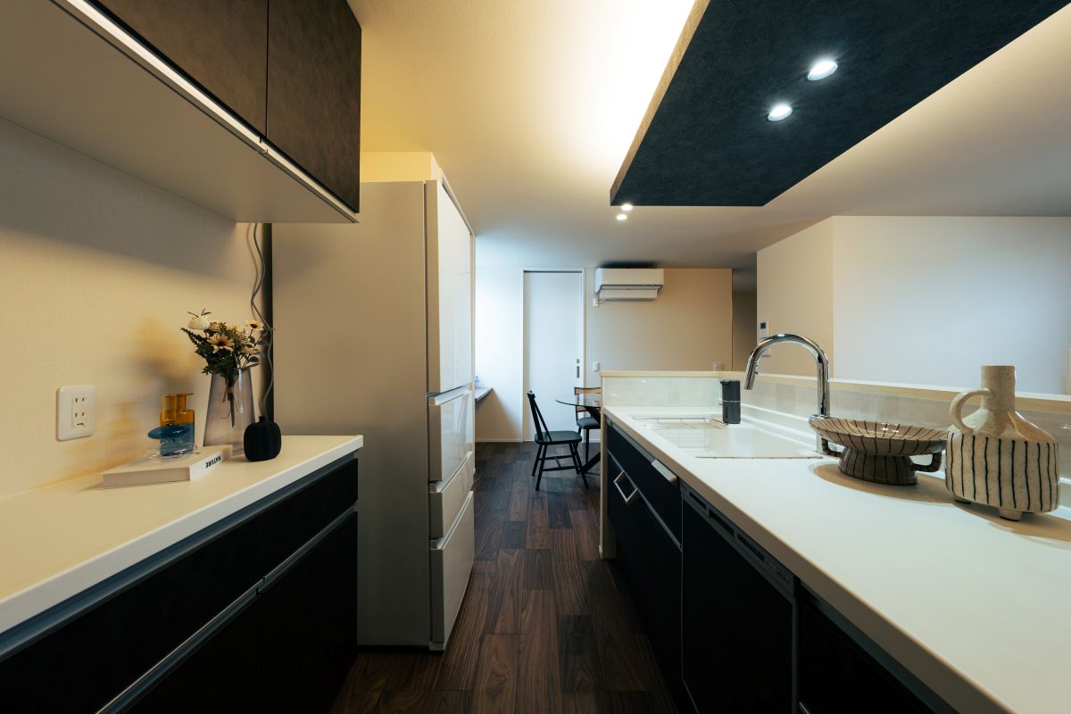 白い天板が特徴的なモダンな対面キッチン。奥に冷蔵庫が置かれ、天井は濃い色の下がり天井になっている。