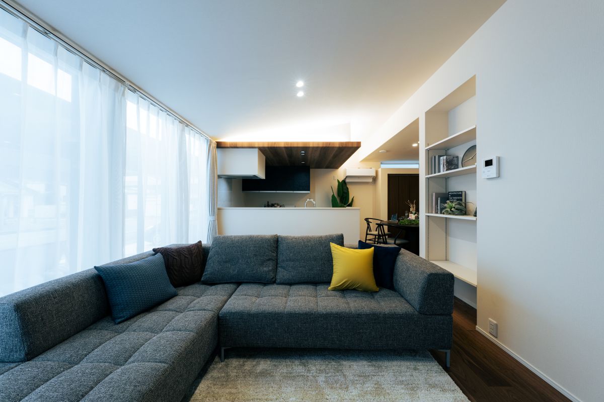 グレーのL字ソファが置かれたリビングルーム。奥にキッチンが見え、壁には白い造作棚が設置されている。
