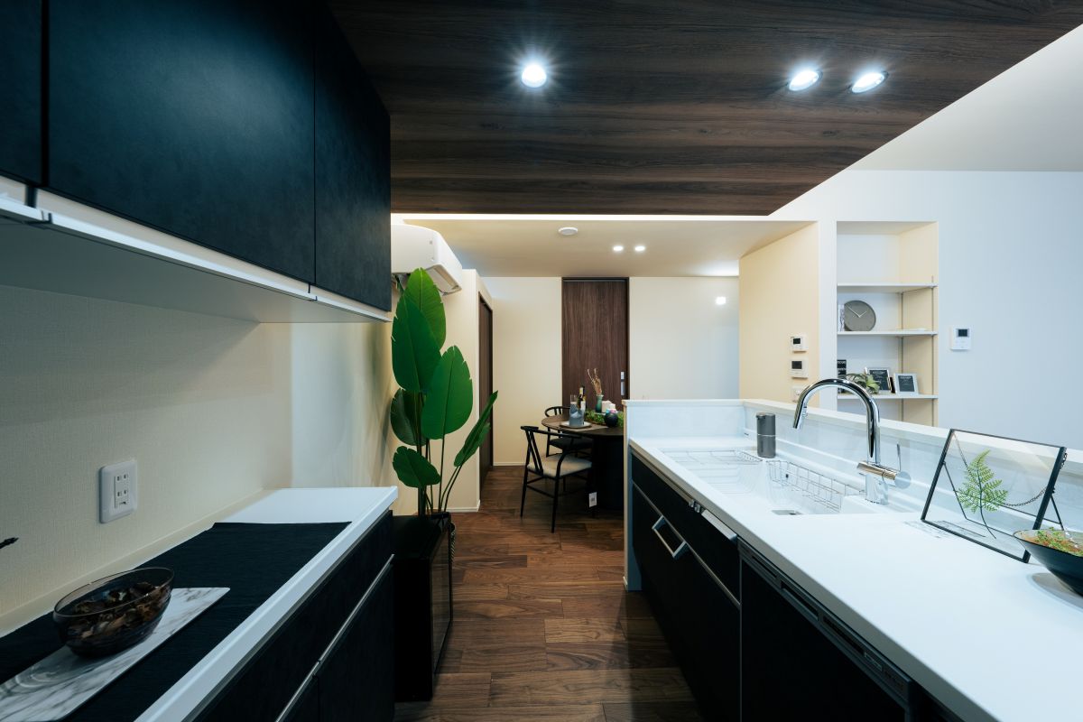 白いシンクカウンターからダイニングを望む。濃い木目調の天井と黒いキャビネットがモダンなキッチン空間。