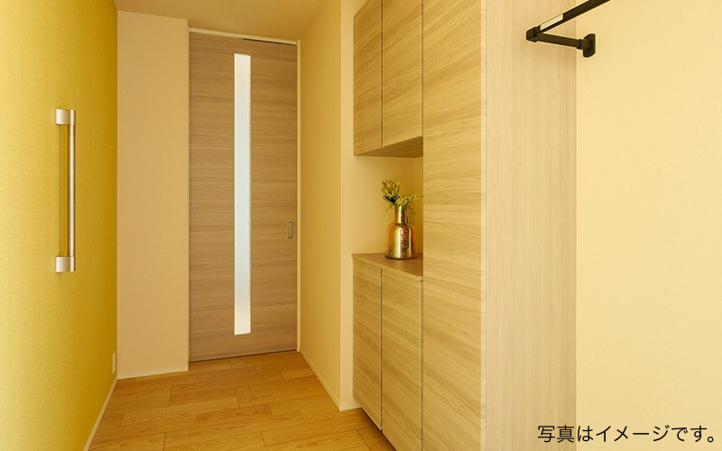 明るい木目調の玄関収納と黄色のアクセントクロスが調和した、温かみのある玄関ホールの内装イメージ。手すりや引き戸も完備されている。