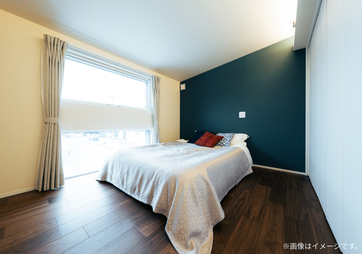 深いブルーのアクセントクロスとダークブラウンの床が落ち着きを演出する寝室。大きな窓から自然光が入り、幾何学模様のベッドカバーが映えるモダンな内装。